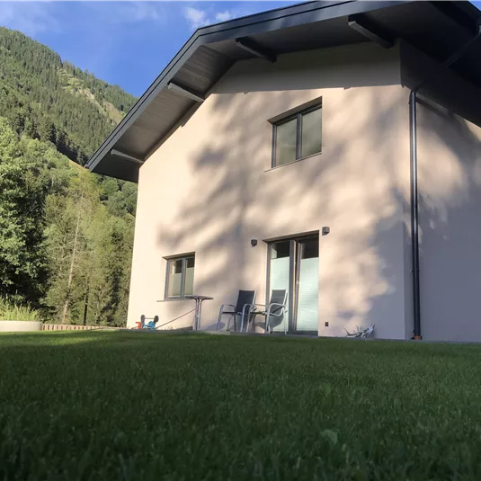 Ein modernes, helles Haus inmitten von grüner Natur. Im Hintergrund sind sanfte Hügel und Bäume zu sehen.