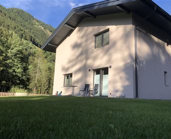 Ein modernes, helles Haus inmitten von grüner Natur. Im Hintergrund sind sanfte Hügel und Bäume zu sehen.