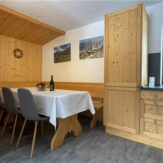 A cozy dining area with a wooden table and chairs. In the background, some landscape pictures are visible on the wall, and a modern kitchen corner can be seen.