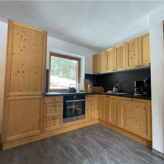 A cozy kitchen with wooden cabinets and modern appliances. Daylight shines through the window, and there is a pleasant seating area.