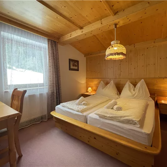 A cozy bedroom with two single beds and wooden furniture. The room has a window with curtains and warm lighting.