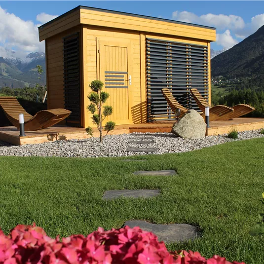 A modern wooden house stands in a well-maintained garden with grass and colorful flowers. In the background, mountains and a blue sky can be seen.