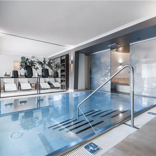 Ein elegantes Hallenbad mit modernem Design und einem klaren Wasser. Neben dem Pool stehen bequeme Liegen und Pflanzen für eine entspannende Atmosphäre.