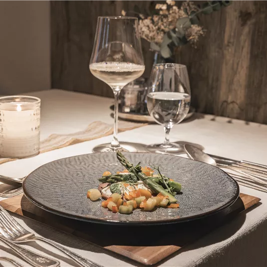 Ein elegant gedeckter Tisch mit einem Hauptgericht auf einem Schwarzgeschirr, garniert mit frischem Gemüse. Im Hintergrund stehen Gläser mit Wein und Wasser sowie eine Kerze für gemütliche Beleuchtung.