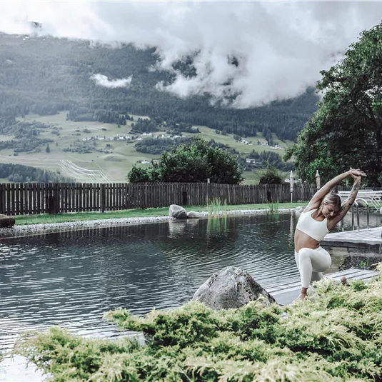 Eine Frau führt eine Dehnübung am Rand eines ruhigen Schwimmbeckens durch. Im Hintergrund sind grüne Hügel und bewölkter Himmel zu sehen.
