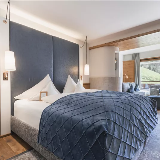 Ein stilvolles Hotelzimmer mit einem großen Bett und einer blauen Bettdecke. Der Raum hat große Fenster mit Blick auf die Natur.