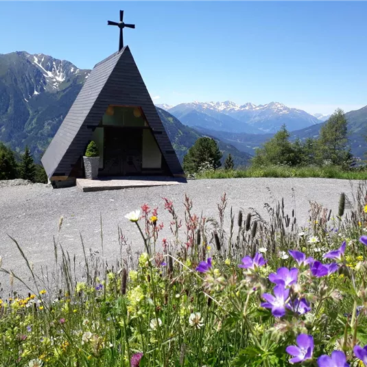 Eine kleine Kapelle mit Kreuz steht auf einem Hügel, umgeben von blühenden Wiesen. Im Hintergrund erheben sich majestätische Berge unter einem klaren blauen Himmel.