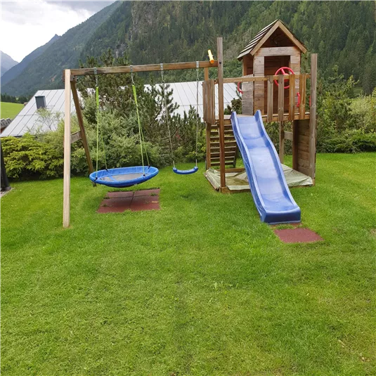 Ein Spielplatz im Grünen mit einer Rutsche, einer Schaukel und einer Nestschaukel. Im Hintergrund sind sanfte Hügel und Bäume zu sehen.