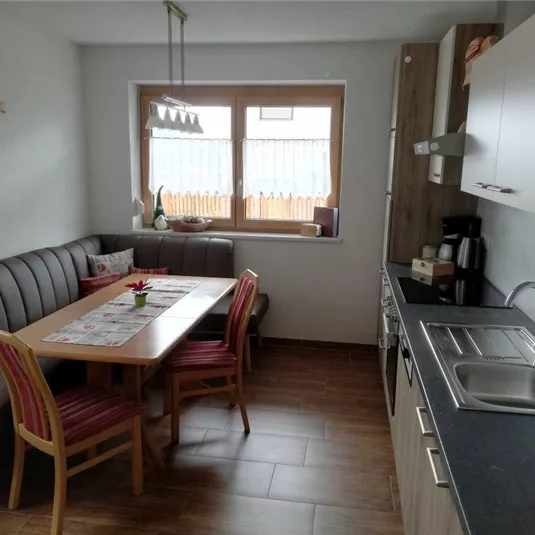 A modern kitchen with a dining area and a cozy seating corner. The room is bright and inviting, with a large window that lets in natural light.
