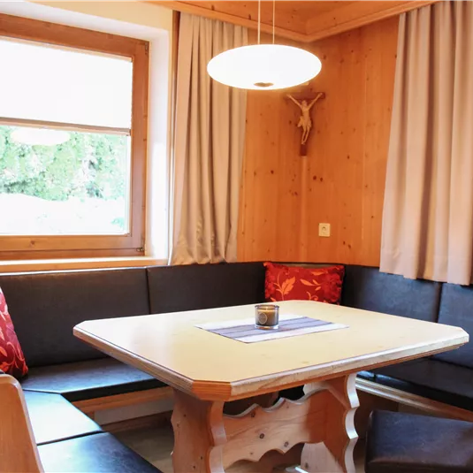 A cozy dining area with wood paneling and a large seating corner. The table is decorated with a subtle tablecloth and a candle, and the window offers a view of greenery.