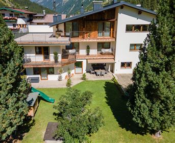 A modern building surrounded by green trees and a well-maintained garden. Outdoor furniture is available on the terrace for cozy hours outside.