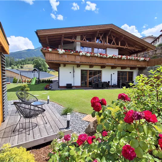 Ein charmantes Haus mit einem großen Balkon und blühenden Blumen im Garten. Im Vordergrund stehen stilvolle Möbel und die schöne Landschaft ist im Hintergrund zu sehen.