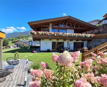 Ein schönes Haus mit Holzverkleidung und Blumenschmuck. Im Vordergrund blühen pinke Hortensien in einem gepflegten Garten.