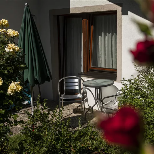 A cozy seating area with a table and chairs in front of a window. Surrounded by blooming roses and green plants.