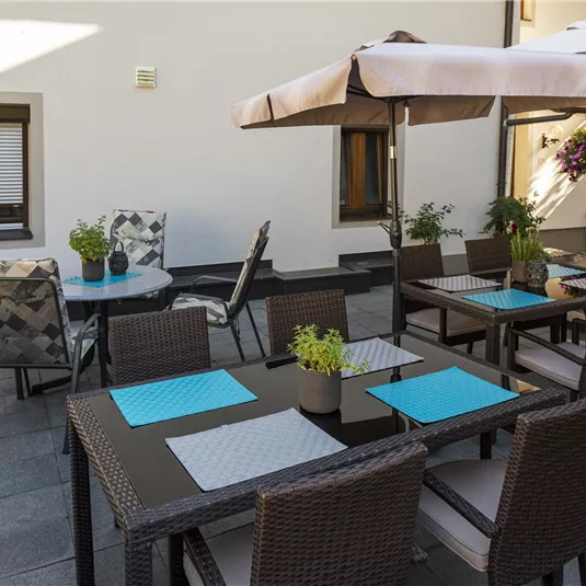A cozy outdoor area with tables and chairs, sheltered by sun umbrellas. Some green plants and decorative table settings create an inviting atmosphere.
