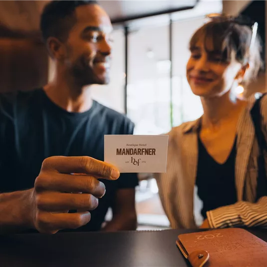 Ein Paar sitzt an einem Tisch und lächelt sich an. Eine Person hält eine Visitenkarte mit dem Namen "Mandarinfahrer" in der Hand.