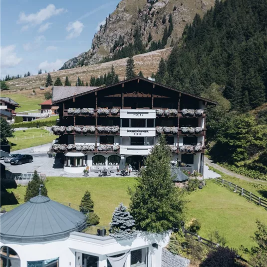 Ein malerisches Hotel inmitten der Alpen, umgeben von Bäumen und Bergen. Im Vordergrund befindet sich ein schöner Poolbereich.