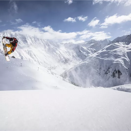 A snowboarder jumps over a snowy slope in the mountains. The sky is clear with some clouds and the scenery is breathtaking.