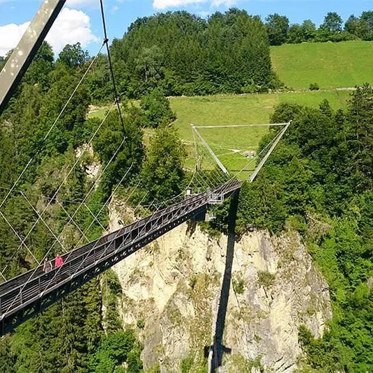 An impressive suspension bridge over a gorge, surrounded by green trees and meadows. In the background, hills and a blue sky are visible.