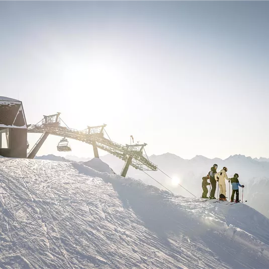A group of skiers stands on a snow-covered slope. In the background, the mountains and a ski lift can be seen.