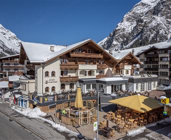 A charming building in alpine style, surrounded by snow-covered mountains. Outside are terraces with sun loungers and yellow sun umbrellas.