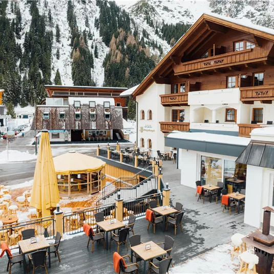 Eine gemütliche Berghütte mit mehreren Terrassen und Holzmöbeln. Im Hintergrund sind schneebedeckte Berge und ein klarer Himmel zu sehen.