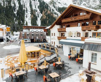 Eine gemütliche Berghütte mit mehreren Terrassen und Holzmöbeln. Im Hintergrund sind schneebedeckte Berge und ein klarer Himmel zu sehen.