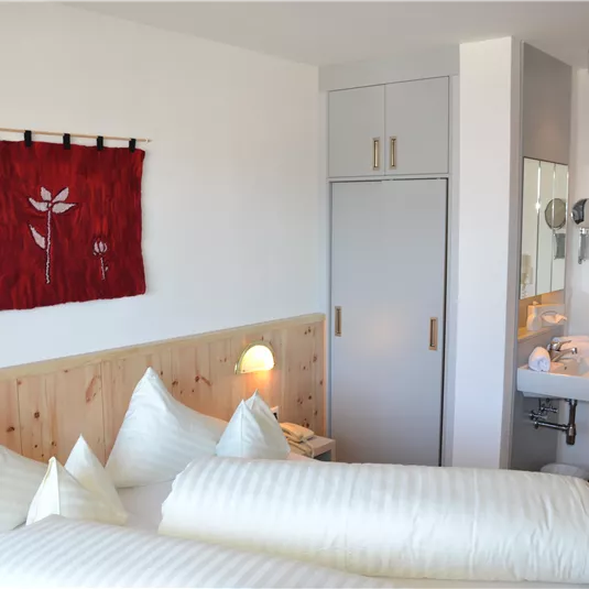 Ein modernes Zimmer mit zwei Betten und dekorativen Kissen. An der Wand hängt ein rotes Wandbild, und es gibt ein Badezimmer mit Waschbecken.
