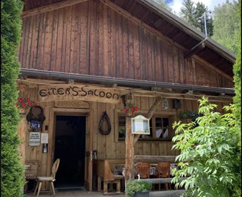 Ein rustikales Holzgebäude mit der Beschriftung "Eiter's Saloon". Vor dem Eingang stehen Tische und Stühle, umgeben von grünen Pflanzen.