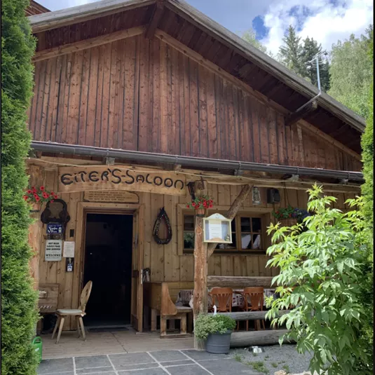 A rustic wooden building with the sign "Eiter's Saloon".
In front of the entrance, there are tables and chairs, surrounded by green plants.