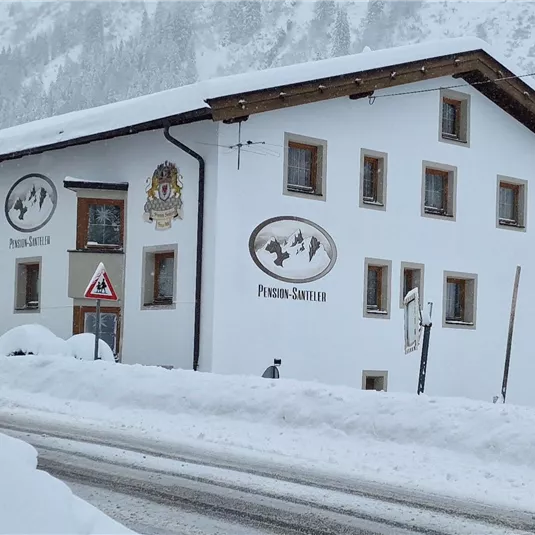 Ein traditionelles Almhüttenhaus im Schnee mit einem schneebedeckten Dach. Die Umgebung zeigt eine ruhige, winterliche Landschaft.
