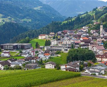 Eine malerische Berglandschaft mit einer kleinen Stadt. Die Häuser liegen in sanften Hügeln, umgeben von grünen Wiesen und bewaldeten Bergen.