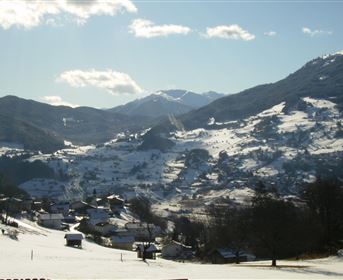 Eine malerische Winterlandschaft mit schneebedeckten Hügeln und kleinen Häusern. Im Hintergrund erheben sich majestätische Berge unter einem klaren blauen Himmel.