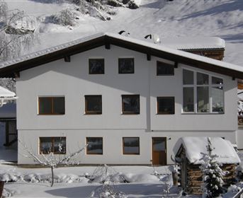Ein modernes Haus im Schnee. Die Umgebung ist von einer winterlichen Landschaft mit Bergen und Bäumen geprägt.
