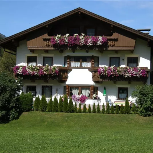 A traditional Alpine house with colorful flower pots. The garden is green and well-maintained, surrounded by mountains.