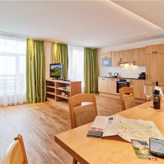 A modern living room with a dining area and a kitchenette. Large windows and green curtains create a bright, inviting atmosphere.