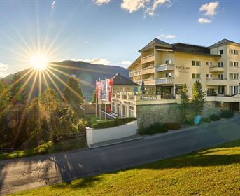 An elegant hotel surrounded by mountains and trees. The sun is shining, giving the scene a warm atmosphere.