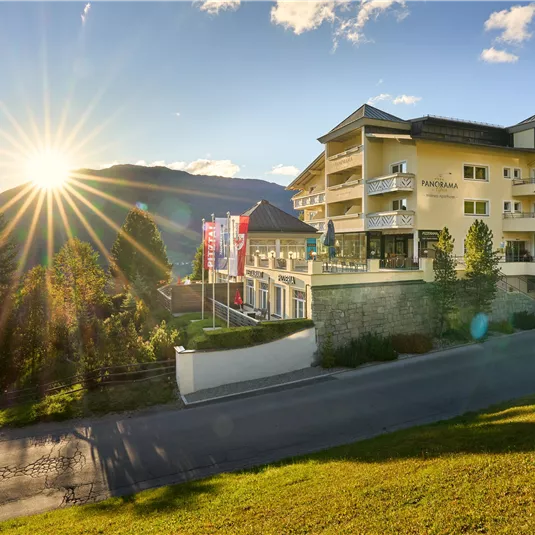 An elegant hotel surrounded by mountains and trees. The sun is shining, giving the scene a warm atmosphere.