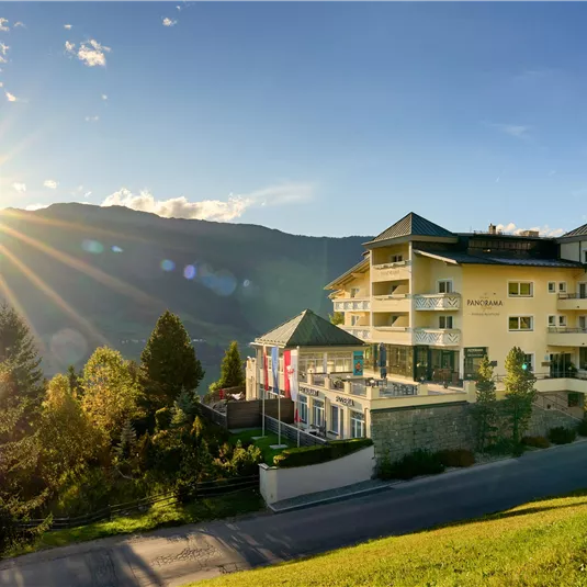 A picturesque mountain landscape with a modern building in the foreground. The sun is shining brightly, creating a warm atmosphere.
