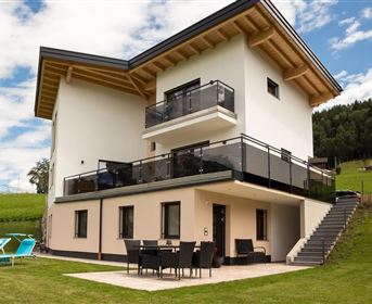 Ein modernes Haus mit Balkon und Terrasse. Im Hintergrund ist eine grüne Wiese und Wald zu sehen.
