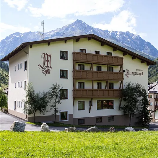 Ein schönes Gebäude mit mehreren Etagen und Balkon im alpinen Stil. Im Hintergrund sind hohe Berge und ein blauer Himmel sichtbar.