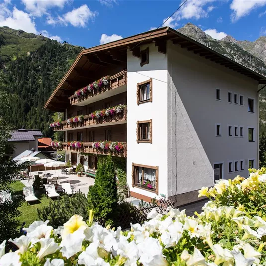 Ein charmantes Hotel mit Balkonen, geschmückt mit Blumen, inmitten einer malerischen Berglandschaft. Der klare Himmel und die grünen Wiesen schaffen eine entspannte Atmosphäre.