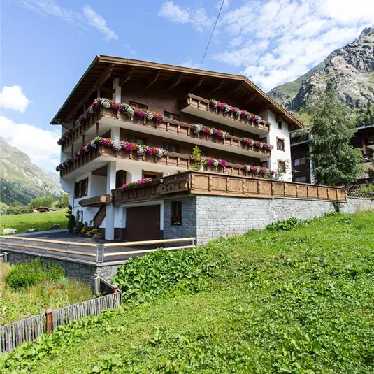 Ein schönes Wohngebäude mit Balkonen und bunten Blumen. Im Hintergrund sind grüne Wiesen und majestätische Berge zu sehen.