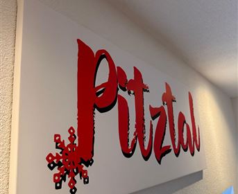Ein Schild mit der Aufschrift "Pitztal" in roter Schrift und einer Schneeflocke. Es ist an einer Wand angebracht und vermittelt eine alpine Atmosphäre.
