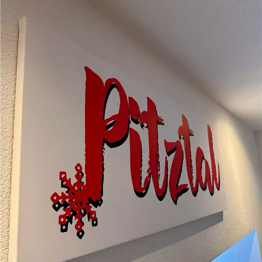 A sign with the inscription "Pitztal" in red letters and a snowflake. It is attached to a wall and conveys an alpine atmosphere.