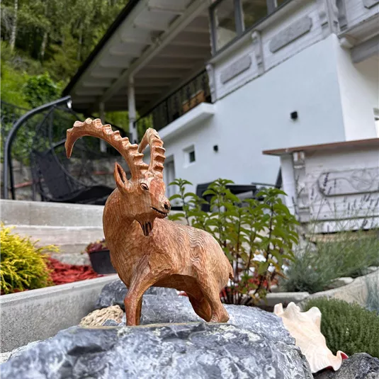 A wooden figure of a ibex stands on a stone in a garden. In the background, a modern house is visible.