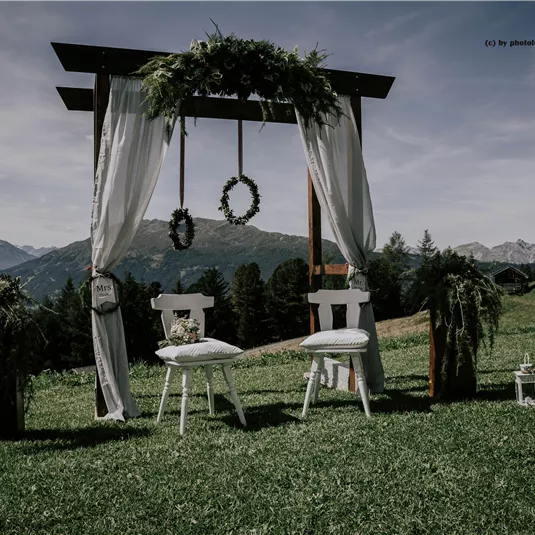 Eine romantische Hochzeitszeremonie im Freien mit einem geschmückten Altar und zwei weißen Stühlen. Der Hintergrund zeigt eine malerische Berglandschaft.