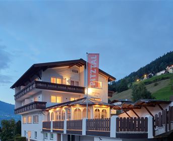A charming hotel in the mountains, illuminated at dusk. Surrounded by green hills and a clear sky.