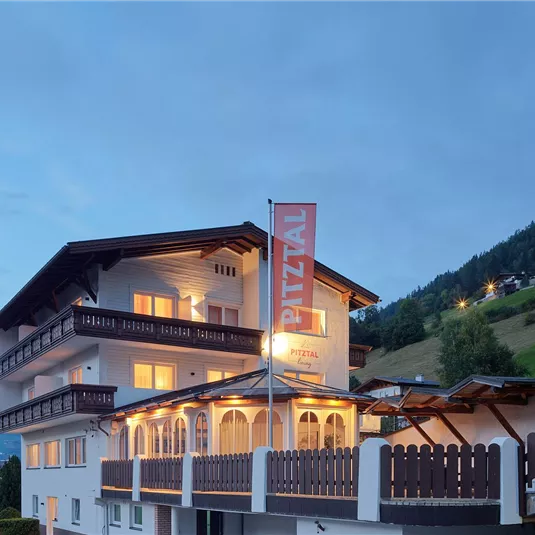 A charming hotel in the mountains, illuminated at dusk. Surrounded by green hills and a clear sky.