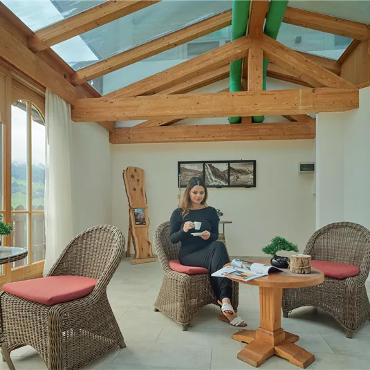 Cozy interior with wooden details and large windows. A person is sitting relaxed with a drink in hand.